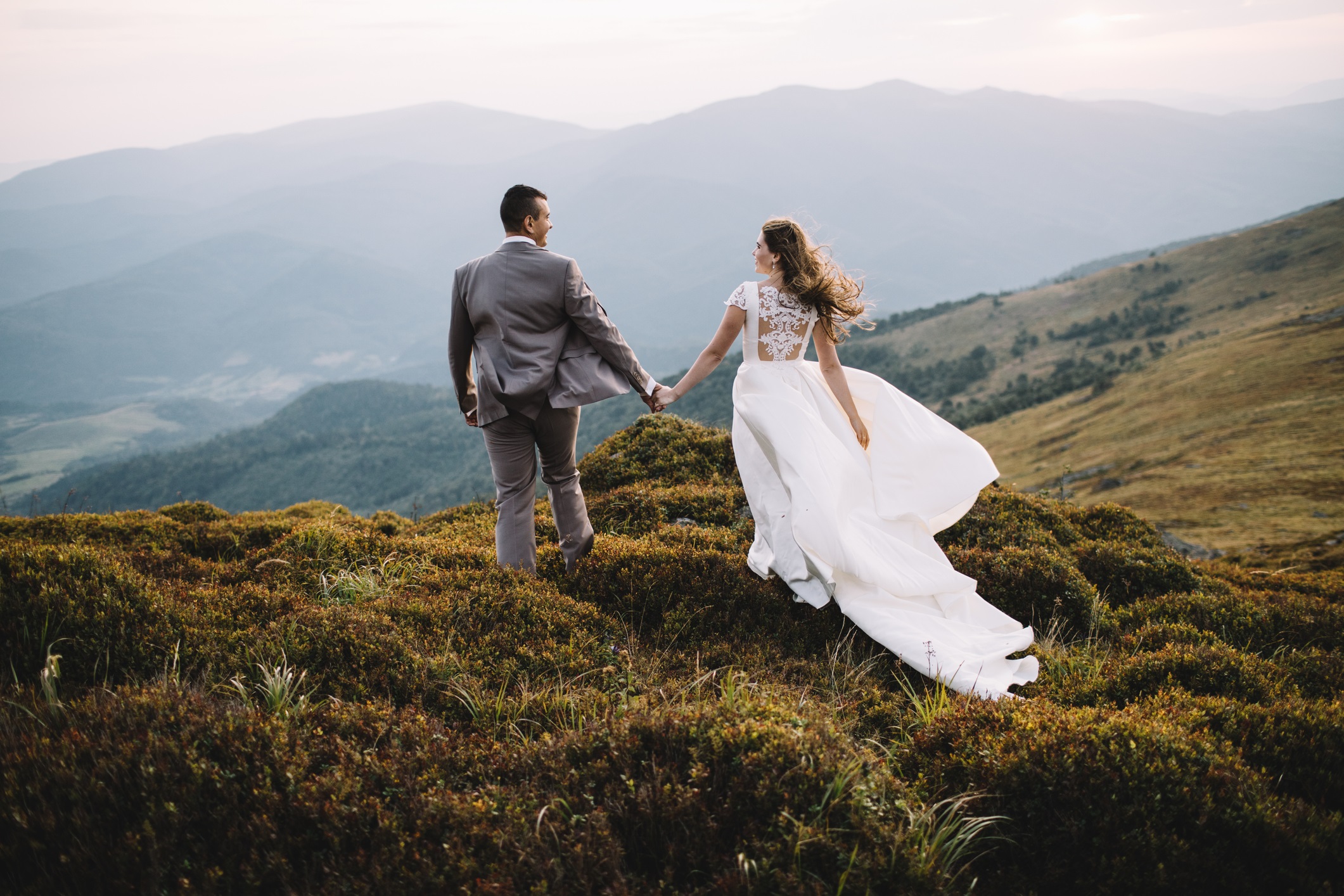 Happy gorgeous bride and groom walking in sun light holding hands, boho wedding couple, luxury ceremony at mountains with amazing view, space for text