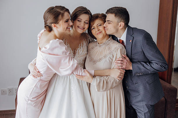 A bride and her three family members are hugging each other. The bride is wearing a white dress and the man is wearing a suit. Scene is warm and loving